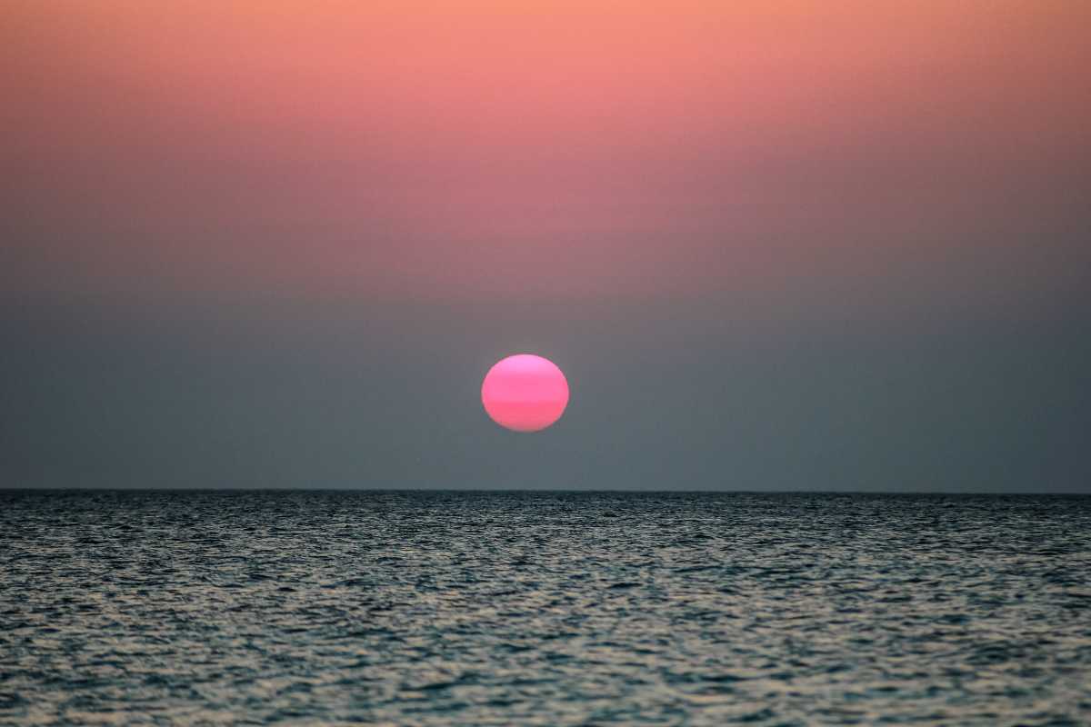 Scena di tramonto sulla spiaggia di Peschici, sabbia dorata, famiglie e coppie sedute, barche dei pescatori sullo sfondo, colori caldi, atmosfera rilassata.
